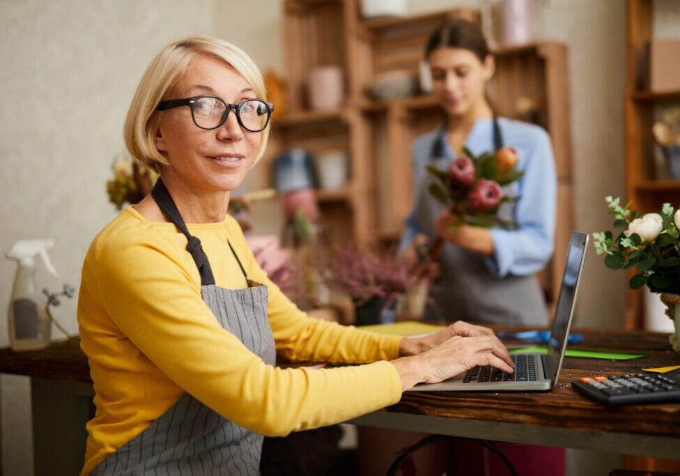 Mature Shop Owner Side view portrait of mature female small business owner using laptop and looking at camera in flower shop, copy space