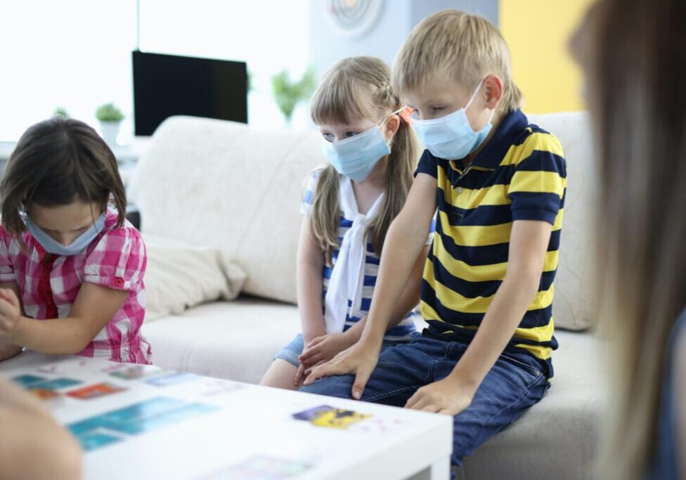 Three children wear protective mask and play board games at home. Playing cards in row on white table.
