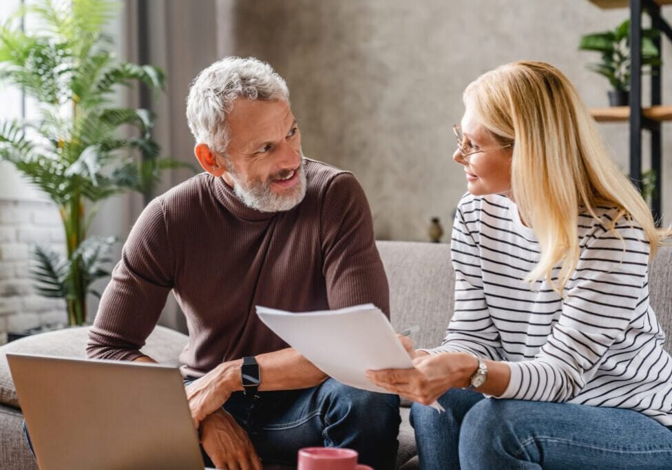 Smiling middle aged couple with laptop studying documents while working on couch at home Smiling middle aged couple with laptop studying documents while working on couch at home