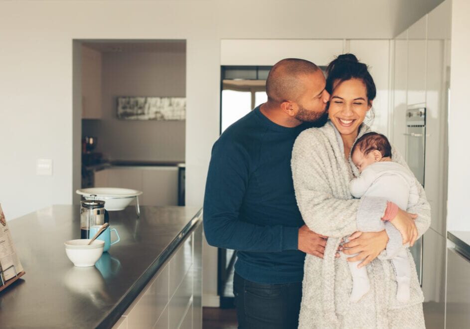 Lovely young family of three in kitchen Man kissing his wife holding a newborn baby boy in kitchen. Lovely young family of three in morning in kitchen.